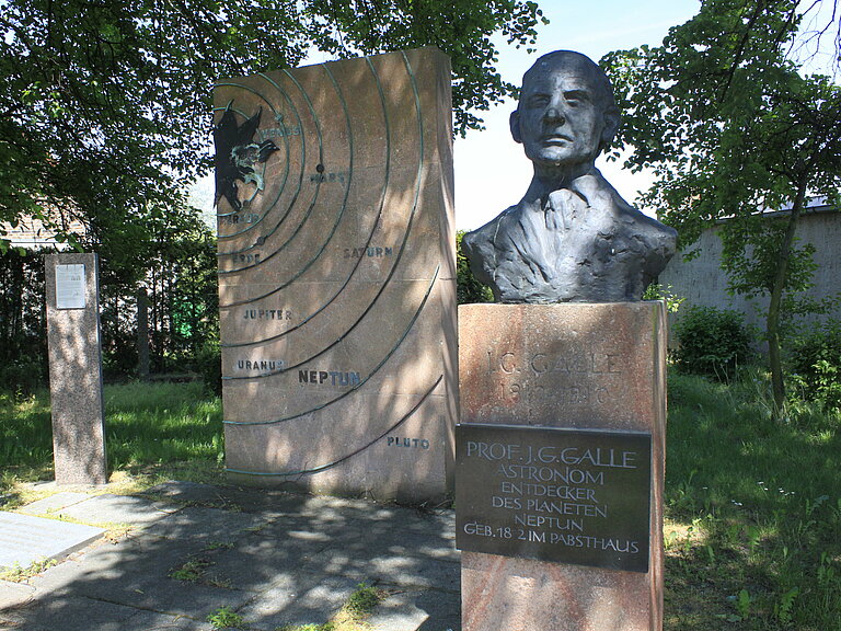 Im Vordergrunud sieht man ein Denkmal von Johann Gottfried Galle. Man sieht sein Gesicht und einen Teil seines Oberkörpers auf einer Stele mit einer Informationstafel. Im Hintergrund sieht man ein Denkmal welches unser Sonnensystem darstellt.