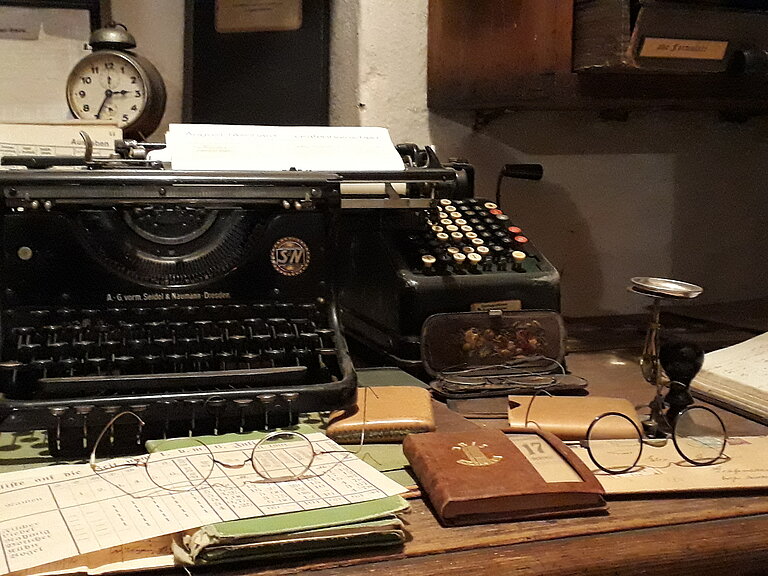Auf diesem Bild sieht man einen Büroarbeitsplatz. Zwei Brillen liegend auf je einem Buch, dahinter sieht man eine alte Schreibmaschine darauf steht ein Wecker.