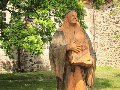 Eine aus Holz geschnitzte Paul-Gerhardt-Skulptur mit einem aufgeschlagenem Buch in den Händen steht vor der Kirchen
