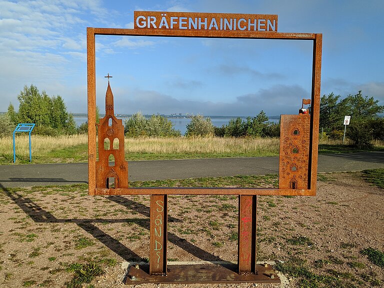 Bilderrahmen aus Stahl mit Schriftzug Gräfenhainichen unterhalb des Stadtbalkons. Im Hintergrund des Bilderrahmens erscheint der Gremminer See.