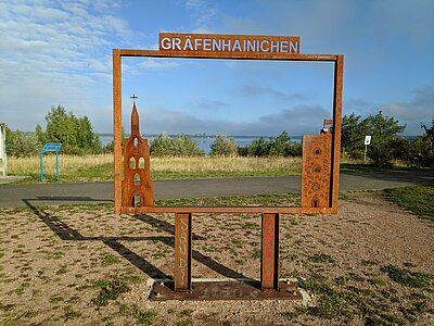 Bilderrahmen aus Stahl mit Schriftzug Gräfenhainichen unterhalb des Stadtbalkons. Im Hintergrund des Bilderrahmens erscheint der Gremminer See.