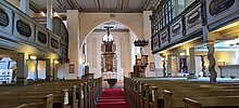 Hier sieht man den Innenraum der Kirche. Rechts und Links sind Sitzbänke zu sehen, außerdem kann man die obere Empore erkennen. Im Hintergrund sieht man den mit schönen Schnitzereien versehenen großen Altar. 