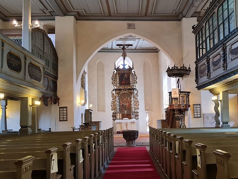 Hier sieht man den Innenraum der Kirche. Rechts und Links sind Sitzbänke zu sehen, außerdem kann man die obere Empore erkennen. Im Hintergrund sieht man den mit schönen Schnitzereien versehenen großen Altar. 