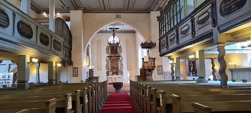 Hier sieht man den Innenraum der Kirche. Rechts und Links sind Sitzbänke zu sehen, außerdem kann man die obere Empore erkennen. Im Hintergrund sieht man den mit schönen Schnitzereien versehenen großen Altar. 