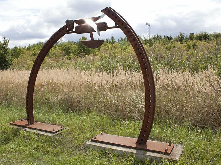 Hier sieht man eine Kunstskulptur, gemacht aus Eisen und Metallen. Es sind zwei gebogene Stangen die in der Mitte leicht zusammenführen und durch eine Waage verbunden sind.