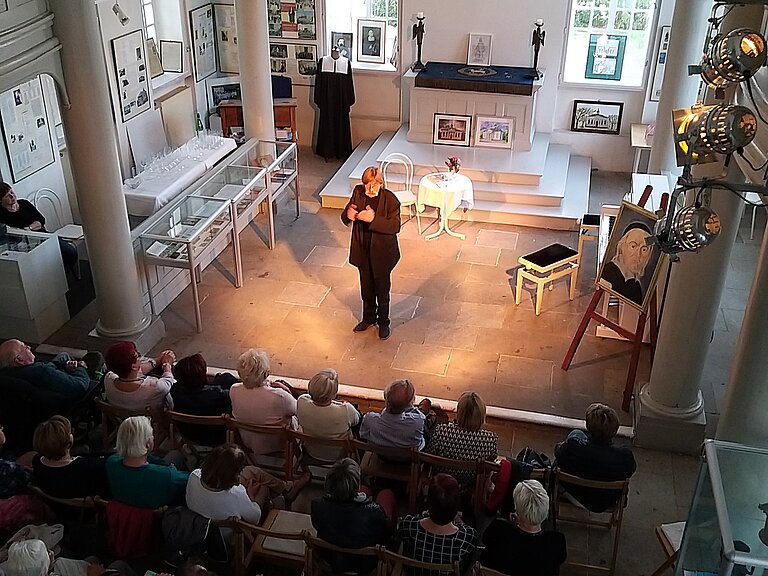 Aufnahme von der Empore in der Paul-Gehardt-Kapelle. Viele Leute hören einem Künstler während seiner Darbietung zu.