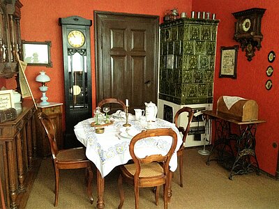 Wohnzimmer mit Standuhr, Kachelofen, Anrichte, gedeckter Tisch und Stühlen um die Jahrhundertwende