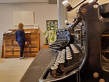 Im Vordergrund sieht man Tasten einer älteren Schreibmaschine. Im Hintergrund ist eine Besucherin zu erkennen, die sich gerade den Setzkastenschrank anschaut.